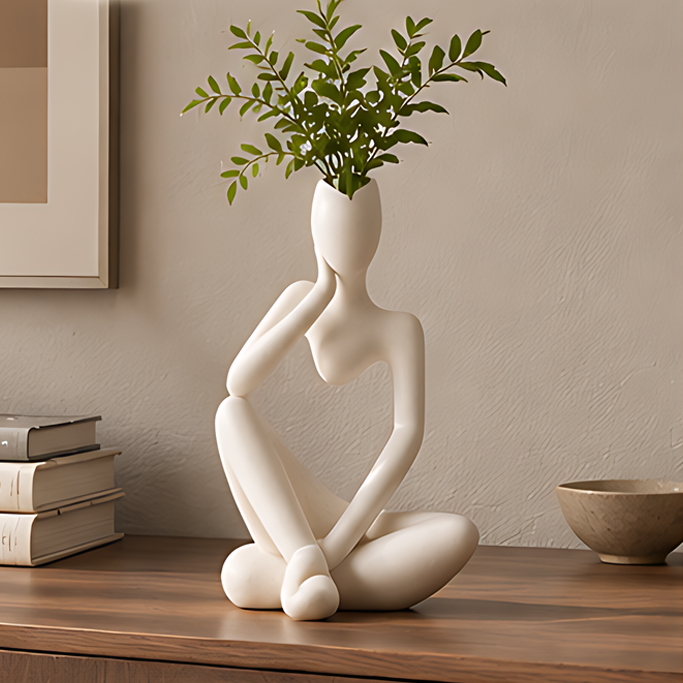 White abstract thinker figure vase with green plant on a wooden desk beside books in a warm interior
